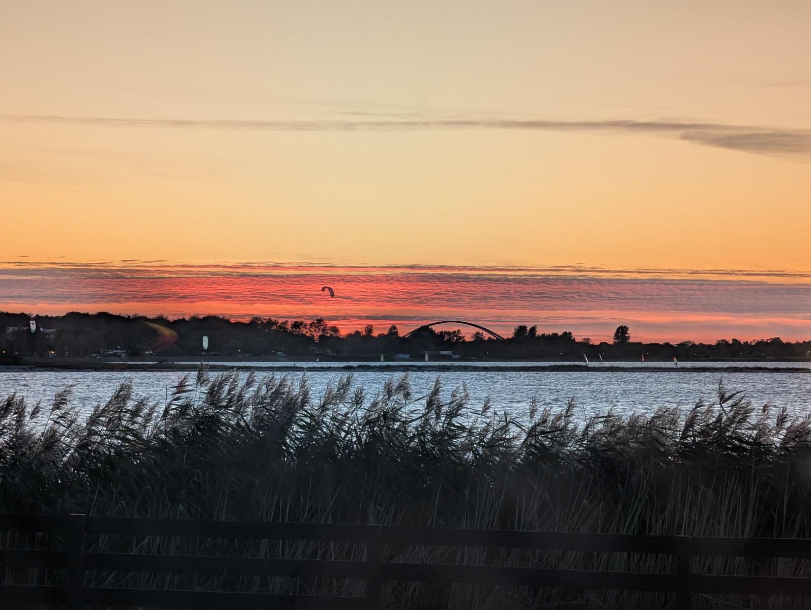 Sonnenuntergang über dem Wasser mit Seglern und einer Brücke im Hintergrund.