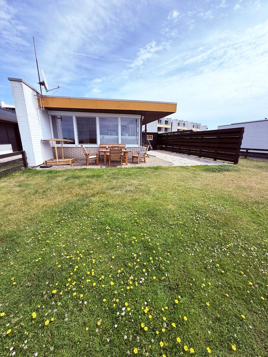 Haus mit Garten, Terrasse und Holztisch mit Stühlen unter blauem Himmel.