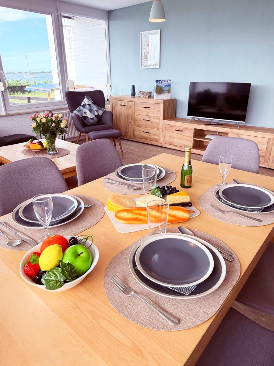 Esstisch mit gedeckten Plätzen, Obst und Käse. Im Hintergrund Wohnzimmer mit TV und Fensterblick.