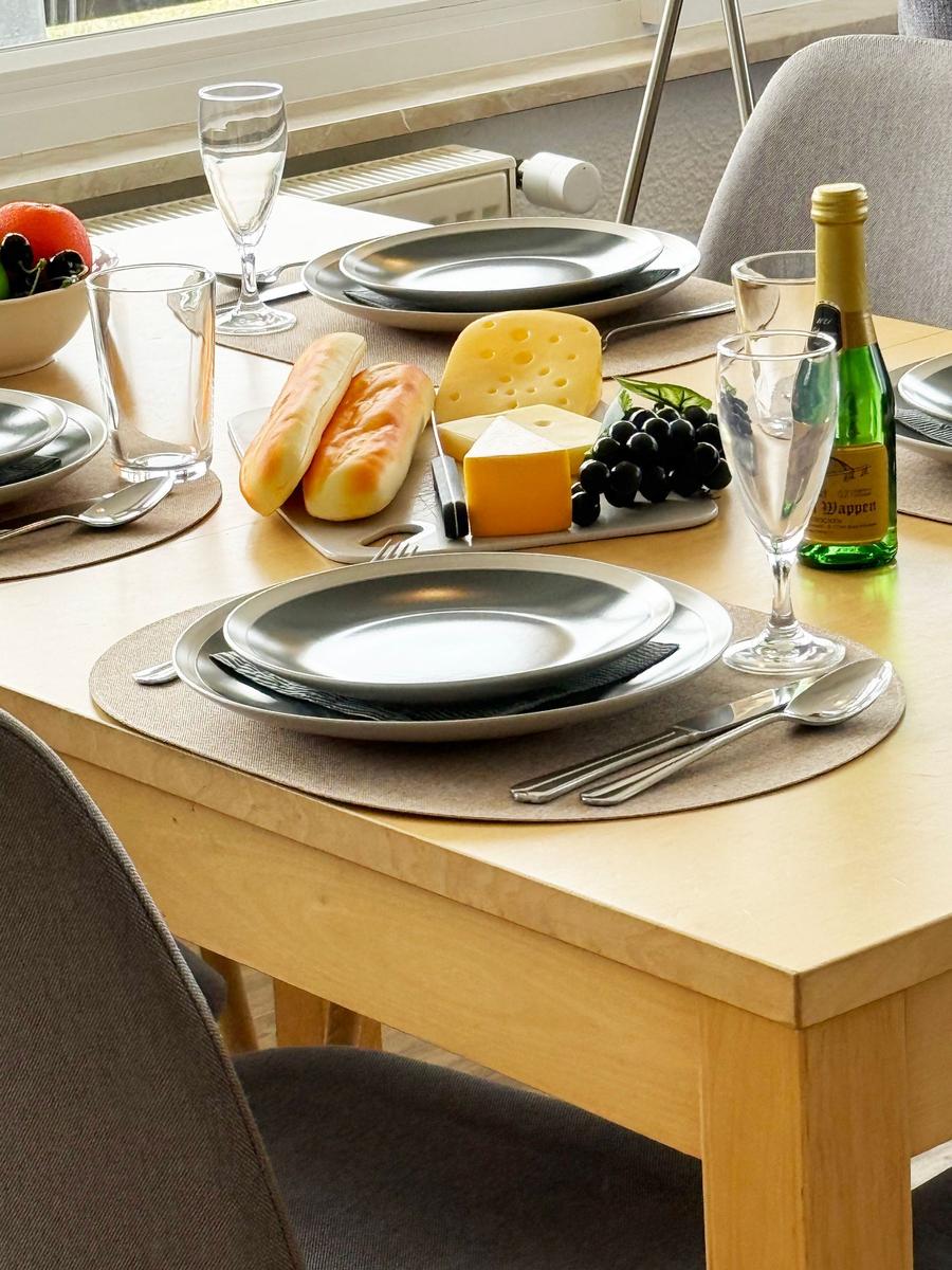 Gedeckter Esstisch mit Platten, Besteck und Käse. Hintergrund: Fenster und Stuhl.