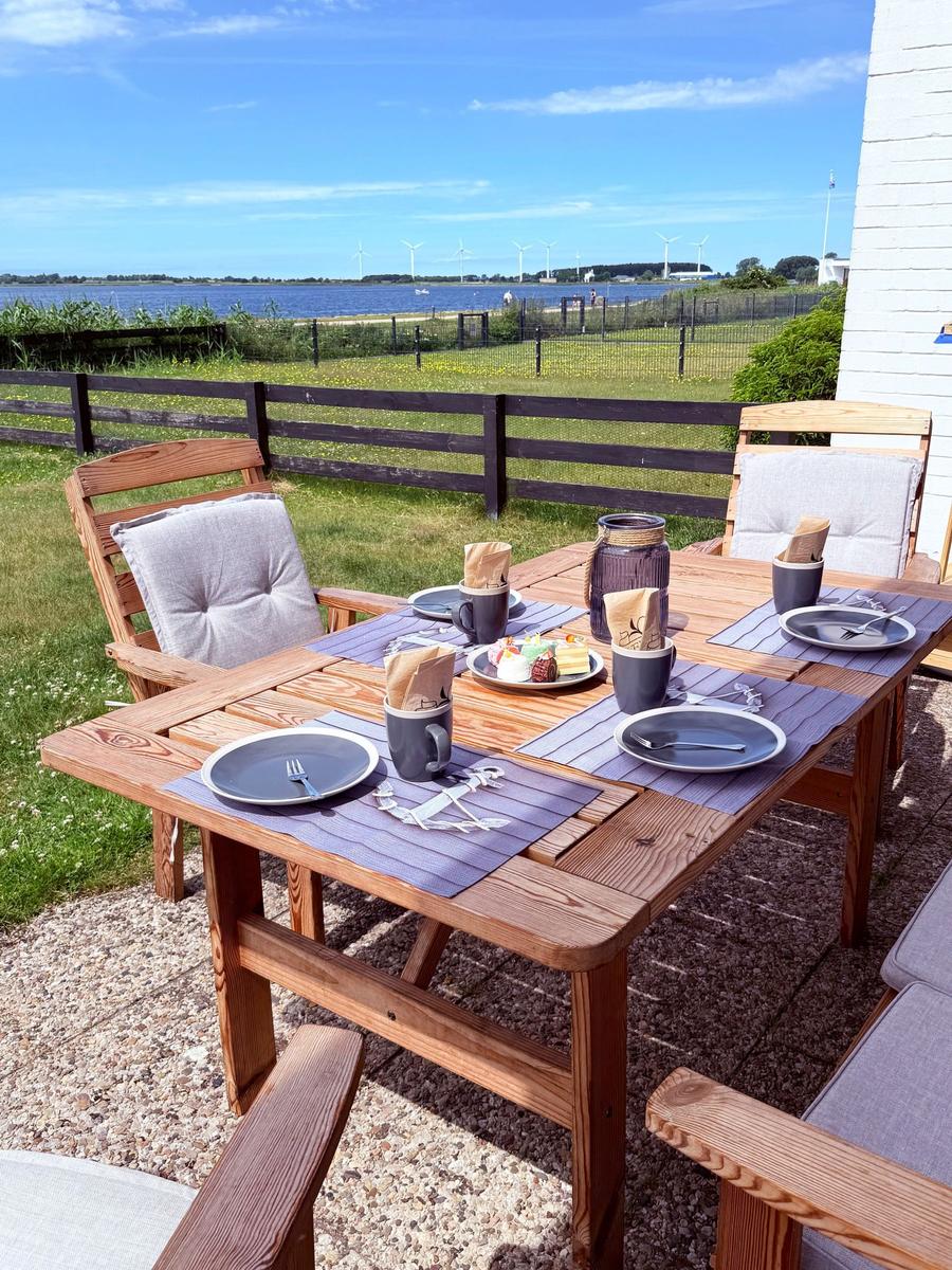 Holztisch mit Stühlen und Tischdecken auf Terrasse mit Blick auf Wasser und Windräder.