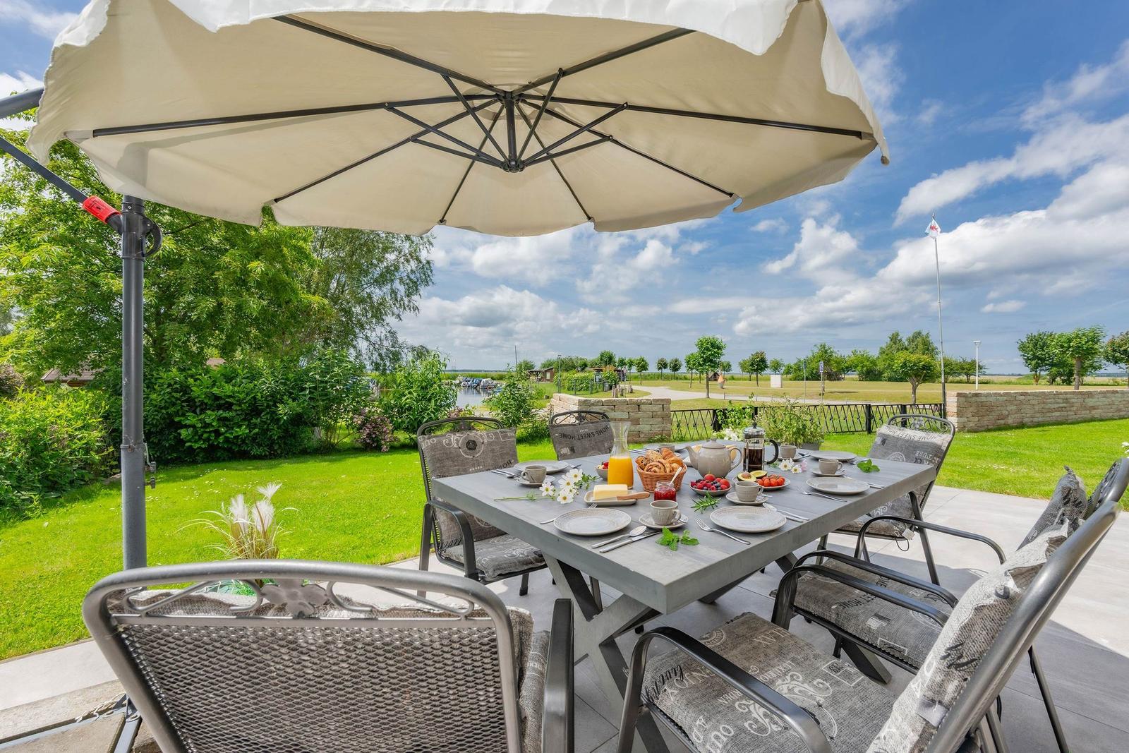 Terrasse mit Tisch, Stühlen und Sonnenschirm. Frühstückszubehör und Blick auf Grünflächen.