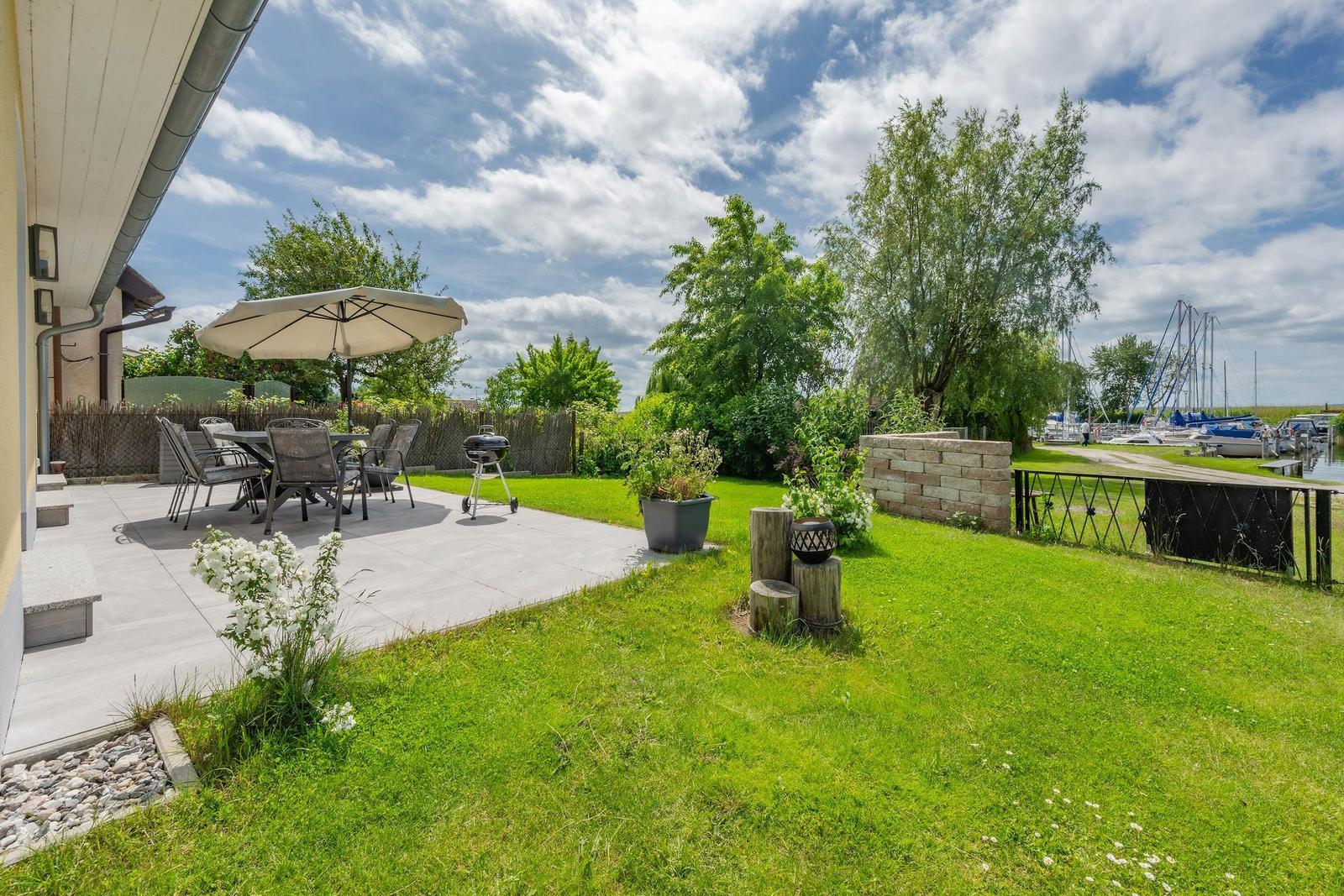 Terrasse mit Tisch, Stühlen und Grill. Grünfläche, Zaun und Bootsanlegestelle im Hintergrund.