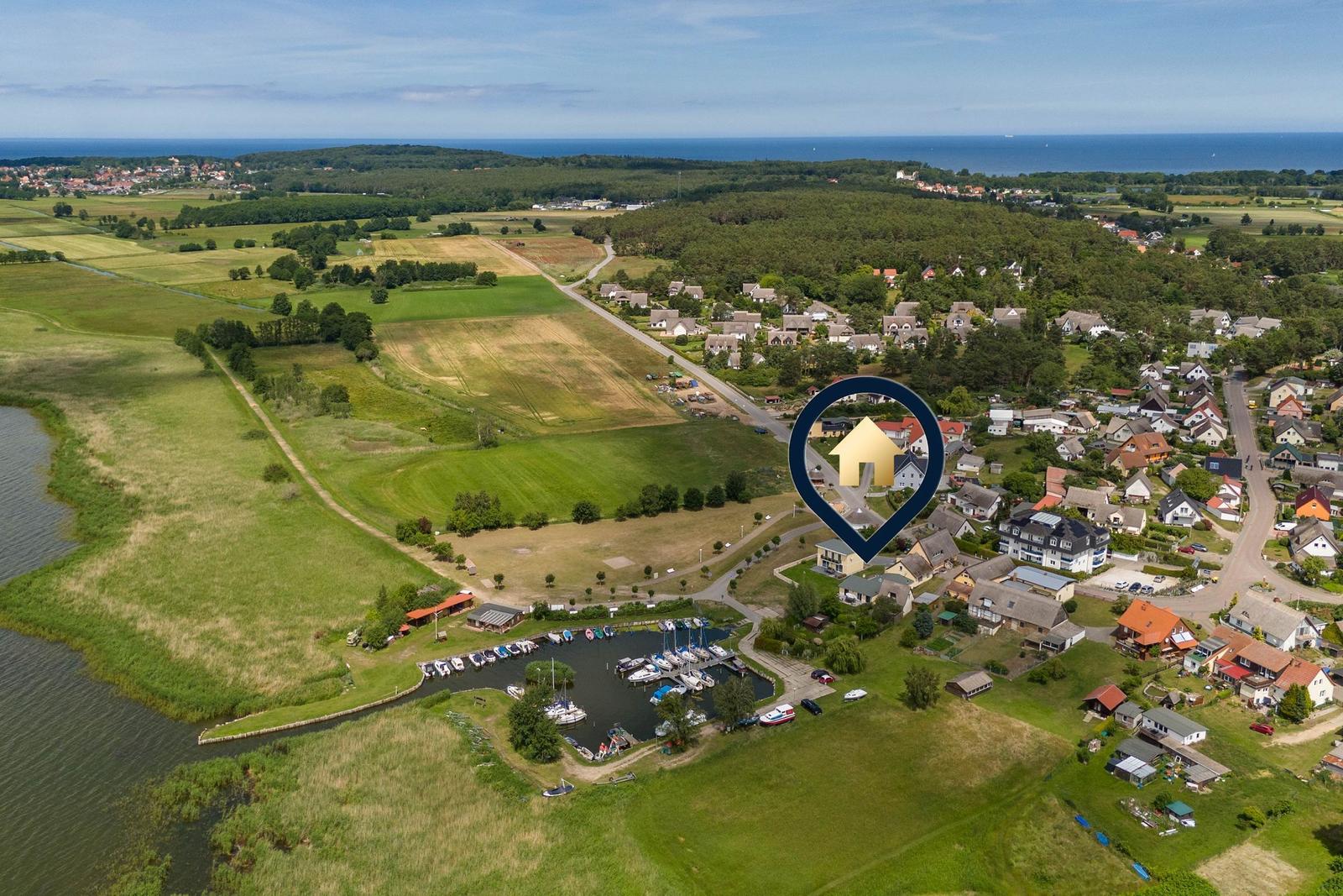Aerielansicht eines Dorfes mit Seeufer, Bootshafen und Wald. Ein markanter Standortpunkt ist hervorgehoben.