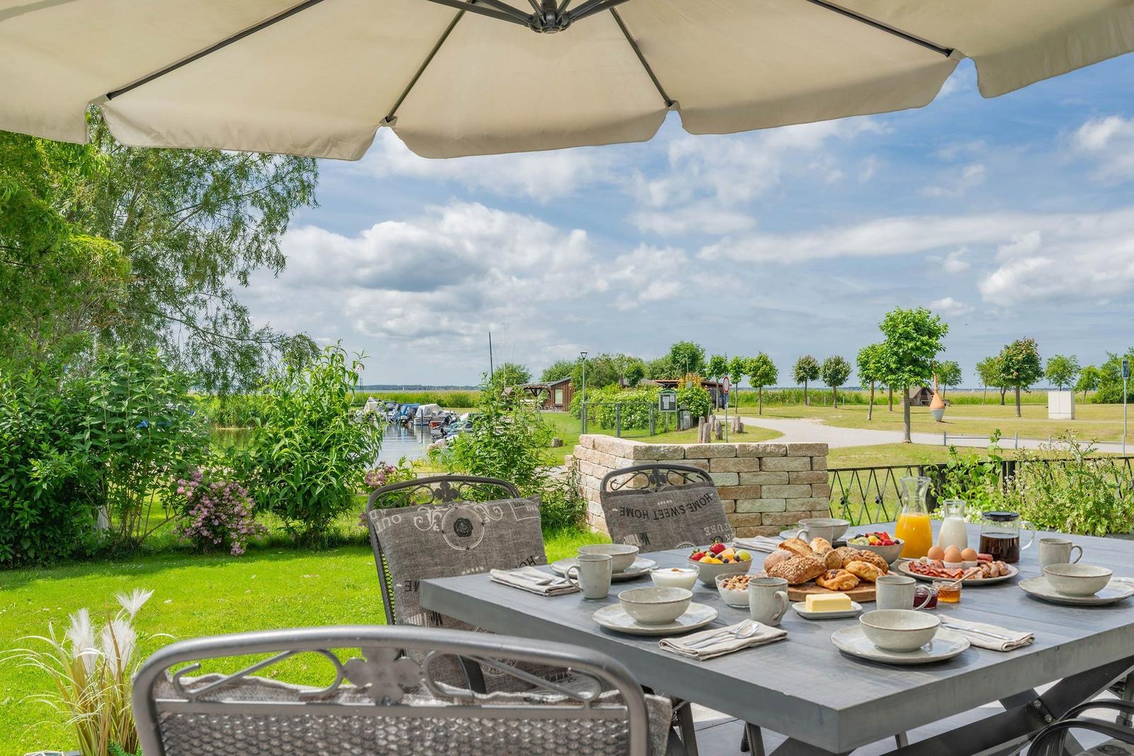 Terrasse mit Tisch, Stühlen und Sonnenschirm. Frühstück auf dem Tisch. Blick auf Wasser und Grün.
