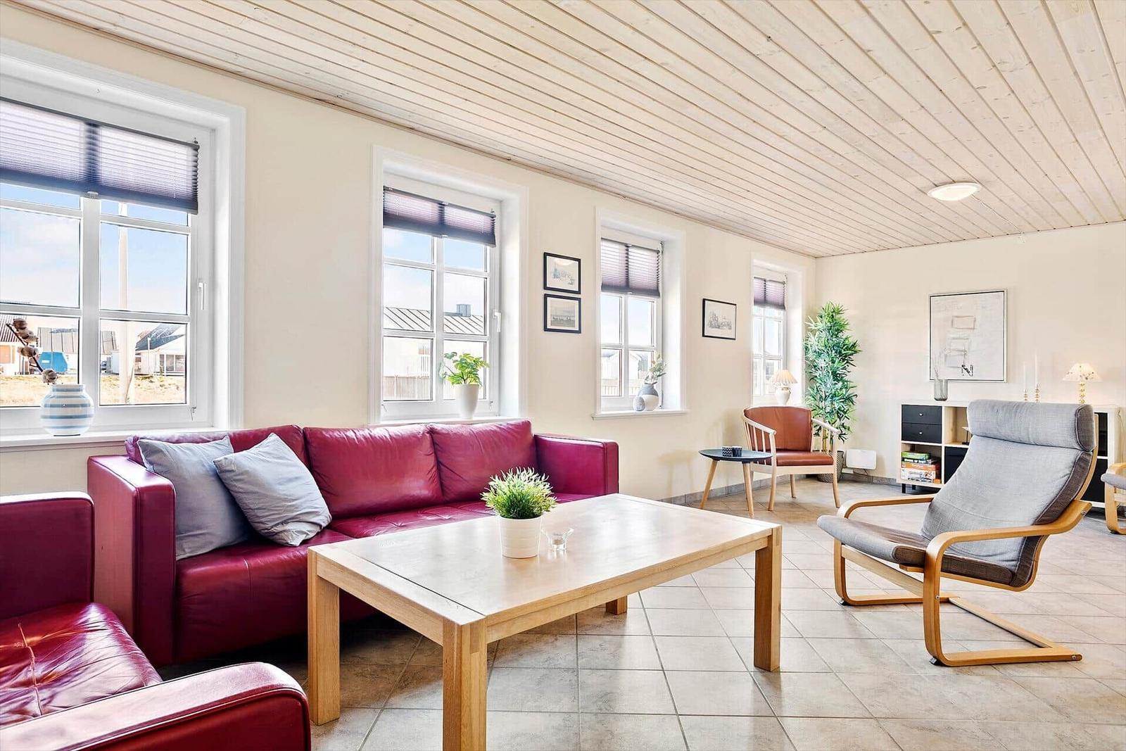 Wohnzimmer mit rotem Sofa, Holztisch und Sessel. Mehrere Fenster und helle Wände.