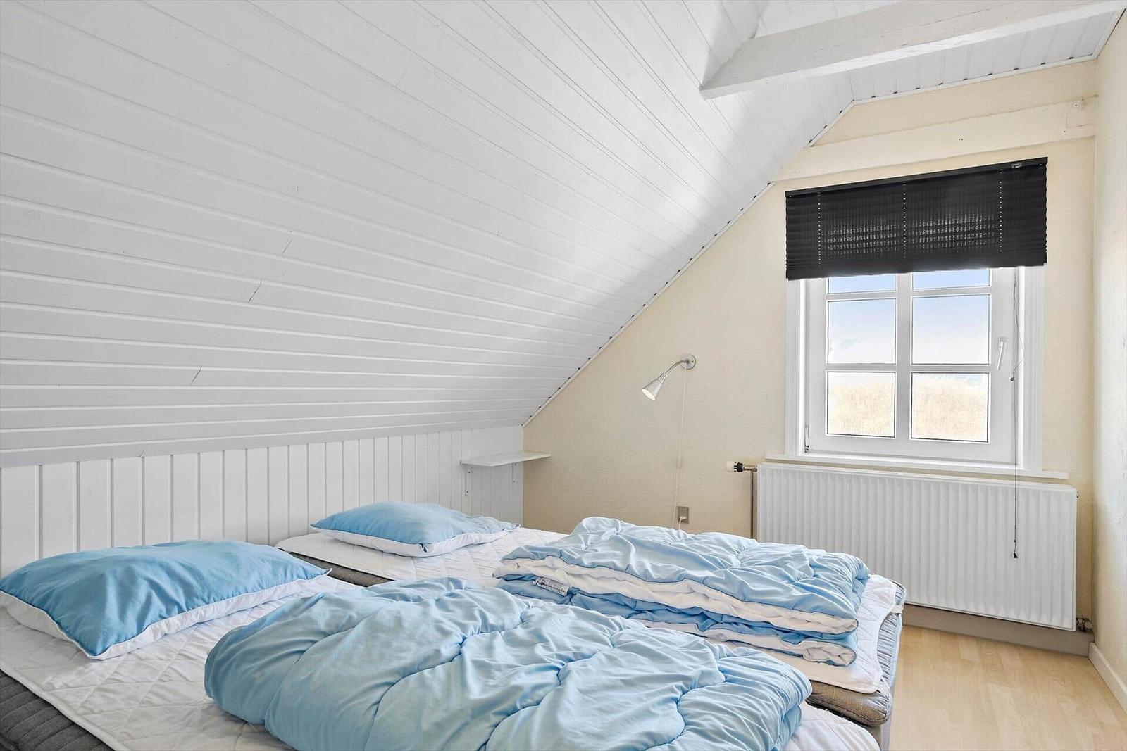 Schlafzimmer mit Dachschrägen, Bett mit blauen Kissen und Decke, Fenster mit Jalousie.