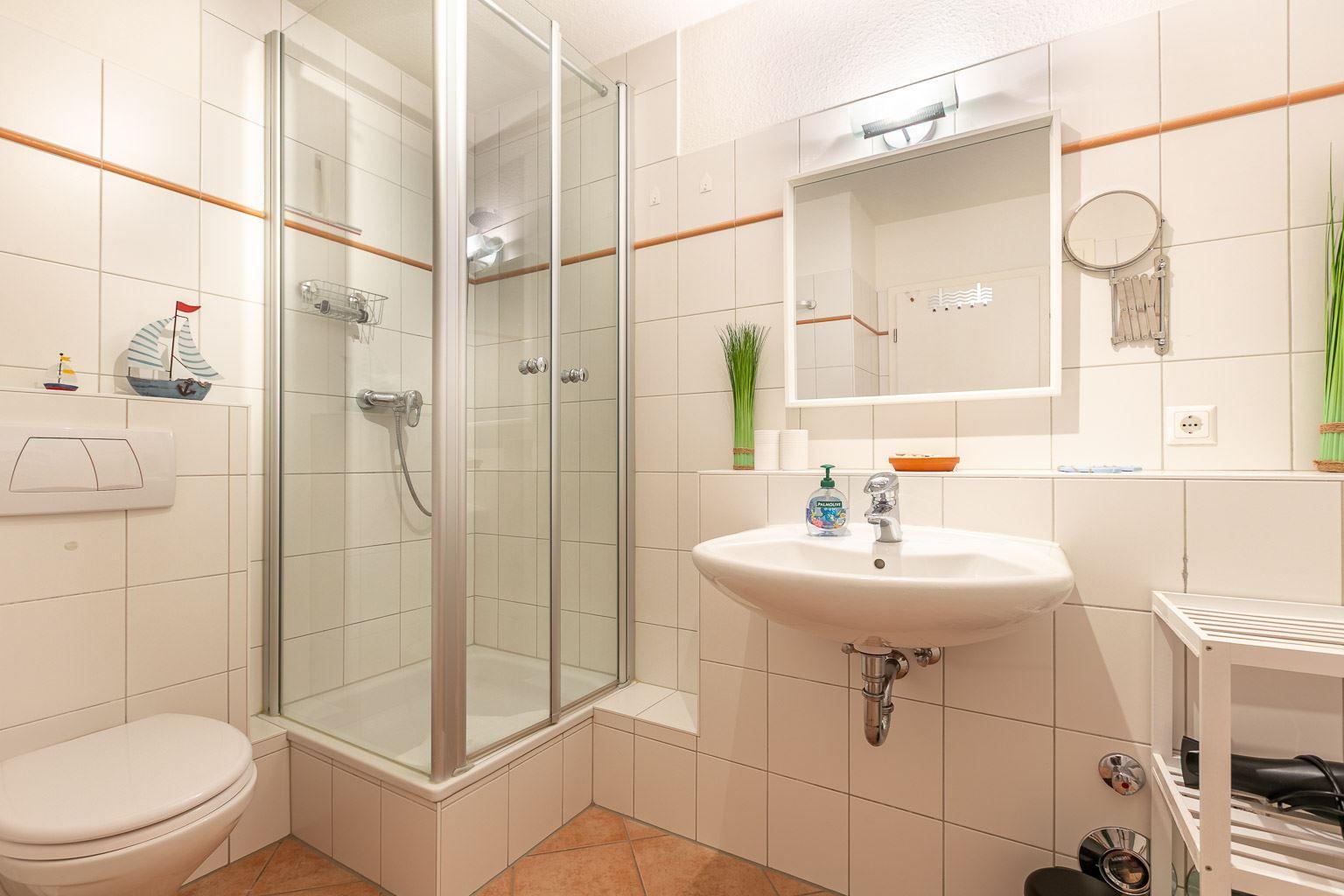Badezimmer mit Dusche, Waschbecken und Toilette. Weiße Fliesen und Holzakzente.
