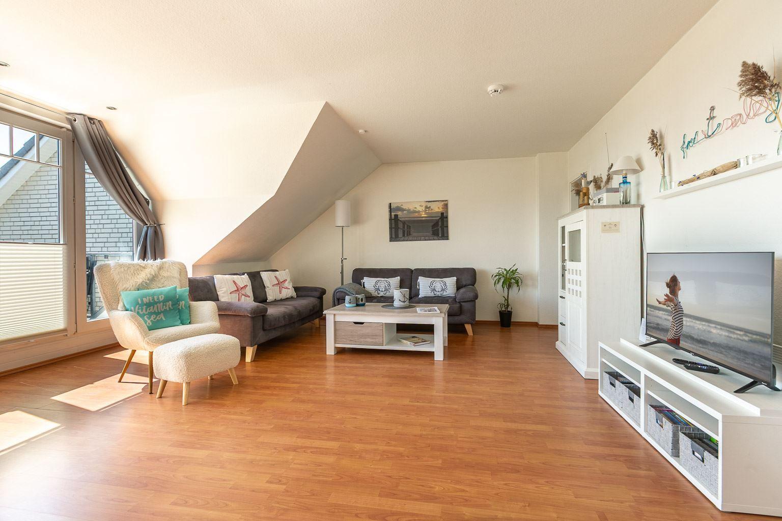 Wohnzimmer mit Sofa, Sessel, Fernseher und Holzboden. Dachschrägen und Fenster sichtbar.