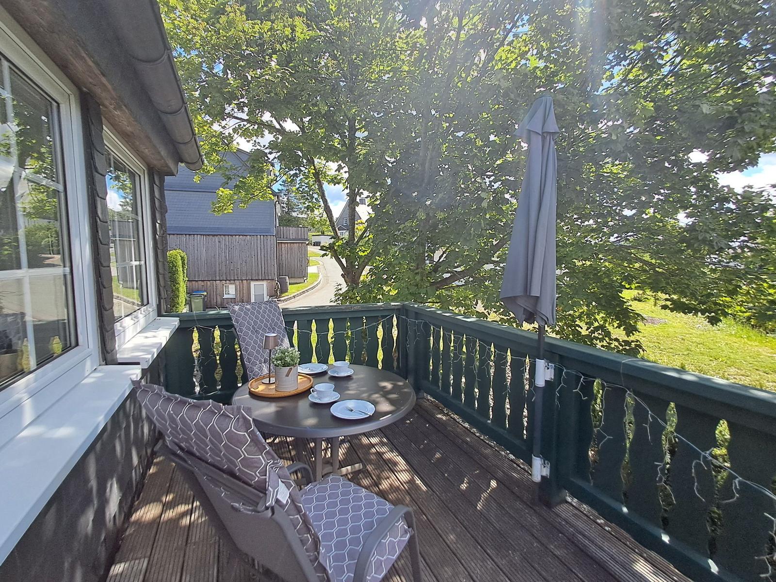Terrasse mit Tisch, Stühlen und Sonnenschirm. Blick auf grüne Bäume und einen Hof.
