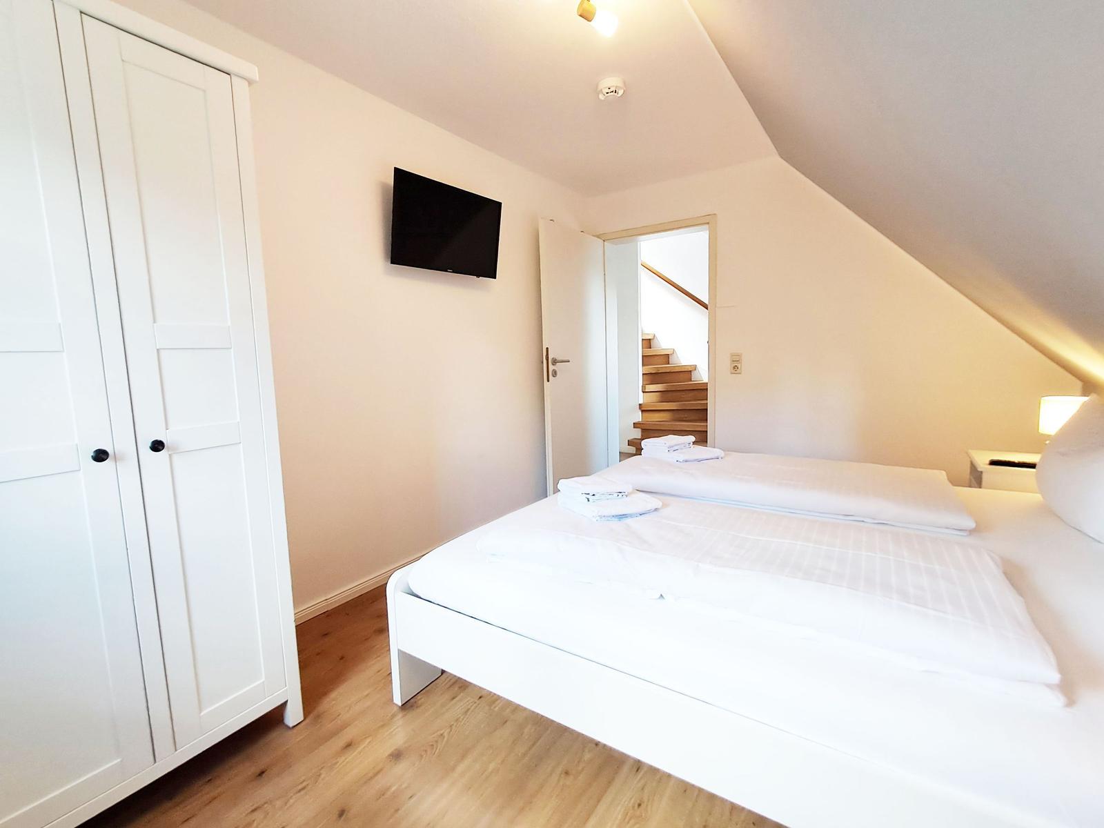 Schlafzimmer mit Doppelbett, Kleiderschrank und TV. Tür führt zu Treppe.