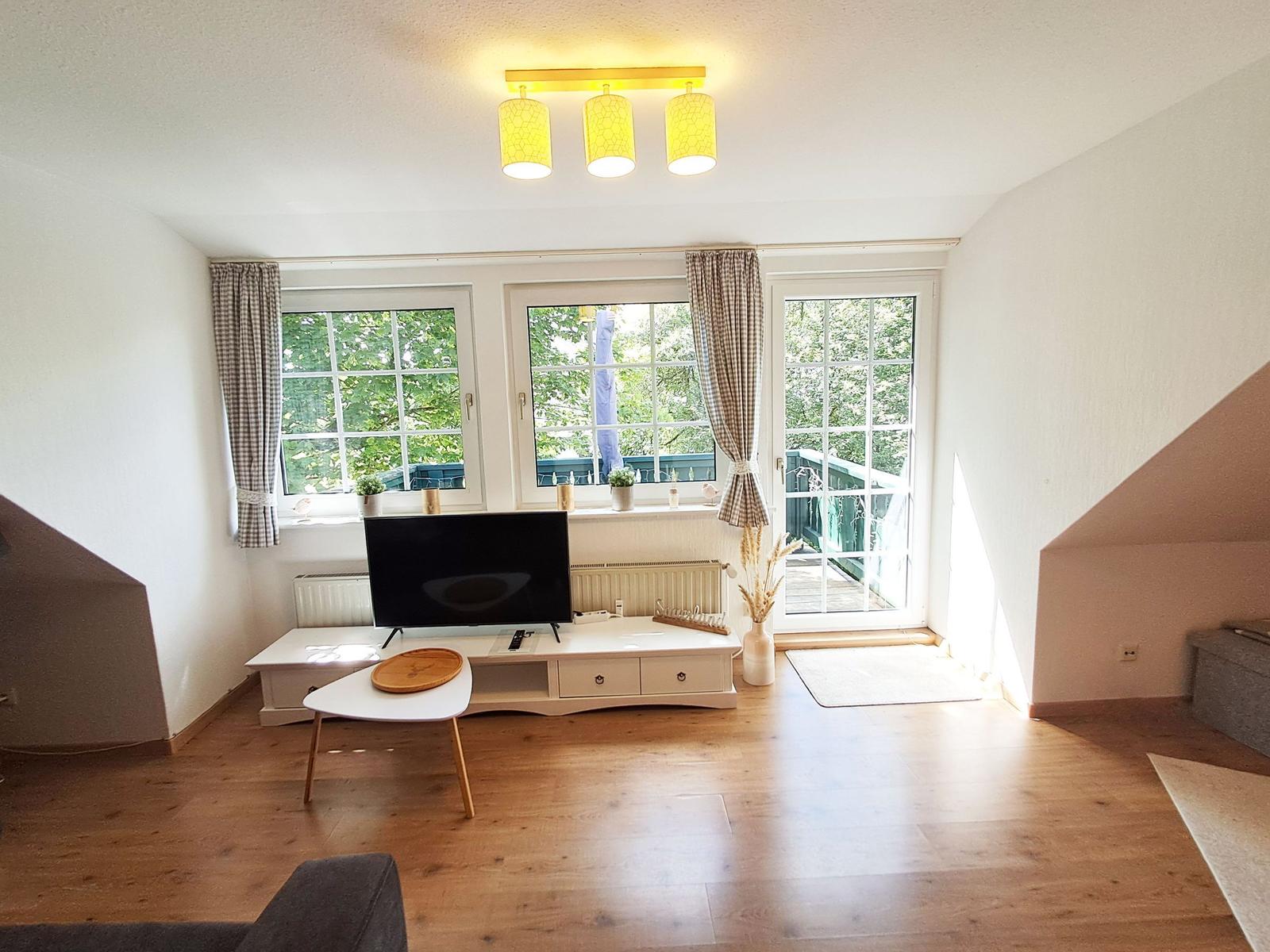 Wohnzimmer mit Holzboden, Fernseher und Fenstern zum Balkon.