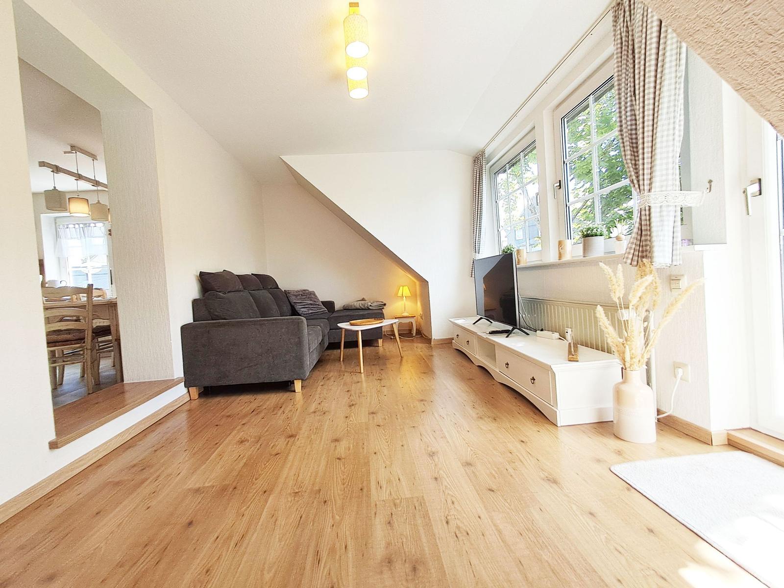 Wohnzimmer mit Sofa, Fernseher und Holzboden. Fenster mit Gardinen. Blick in die Küche.