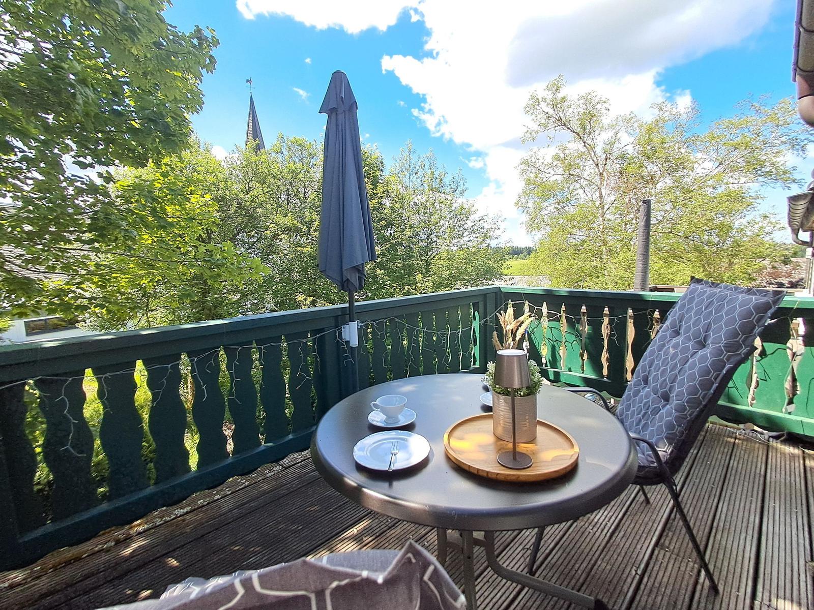 Terrasse mit Tisch, Stuhl und Sonnenschirm. Blick auf grüne Bäume und einen Kirchturm.