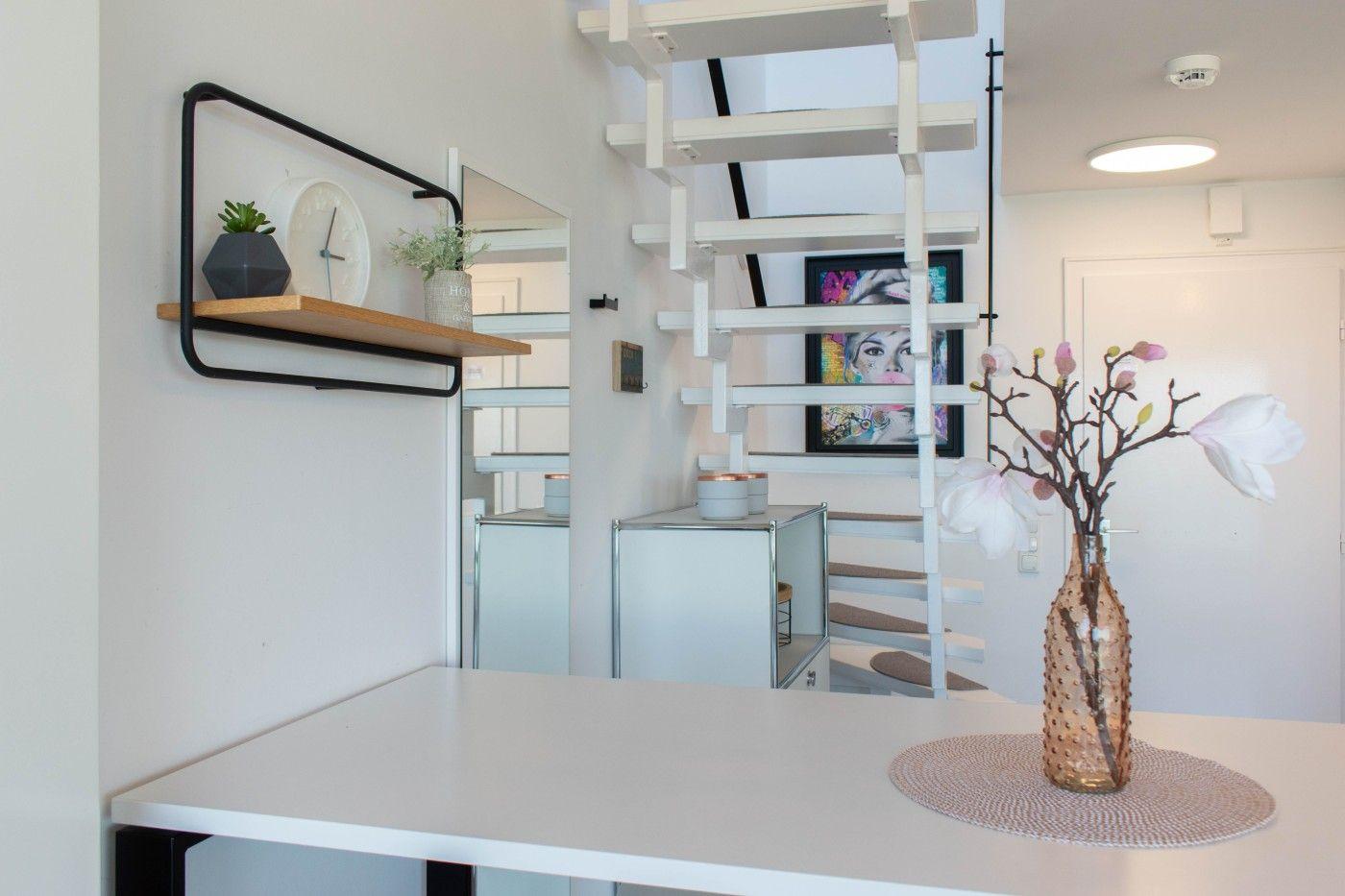 Helle Wohnung mit Treppe, Tisch und Wandregal mit Pflanzen und Uhr.