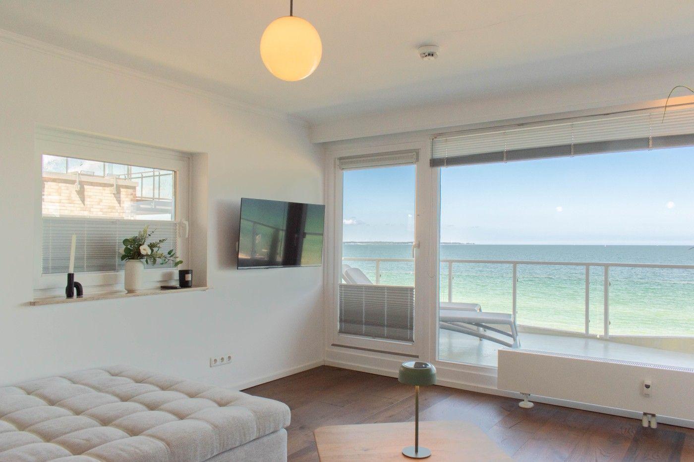 Wohnzimmer mit Meerblick, TV und Balkon. Holzboden und weiß gestrichene Wände.