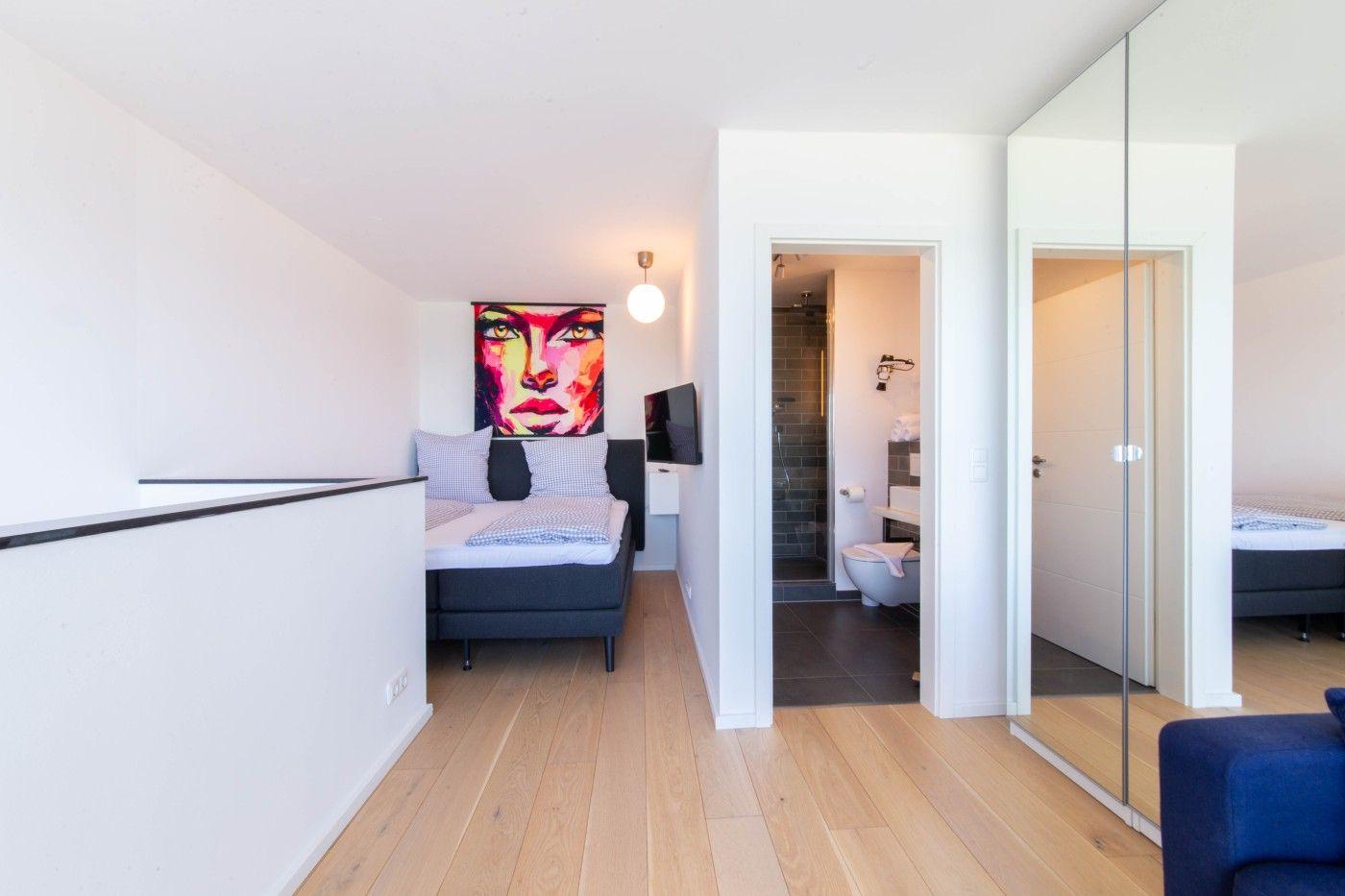 Schlafzimmer mit Bett, Wandbild und Tür zum Bad. Holzboden und Spiegeltür sichtbar.