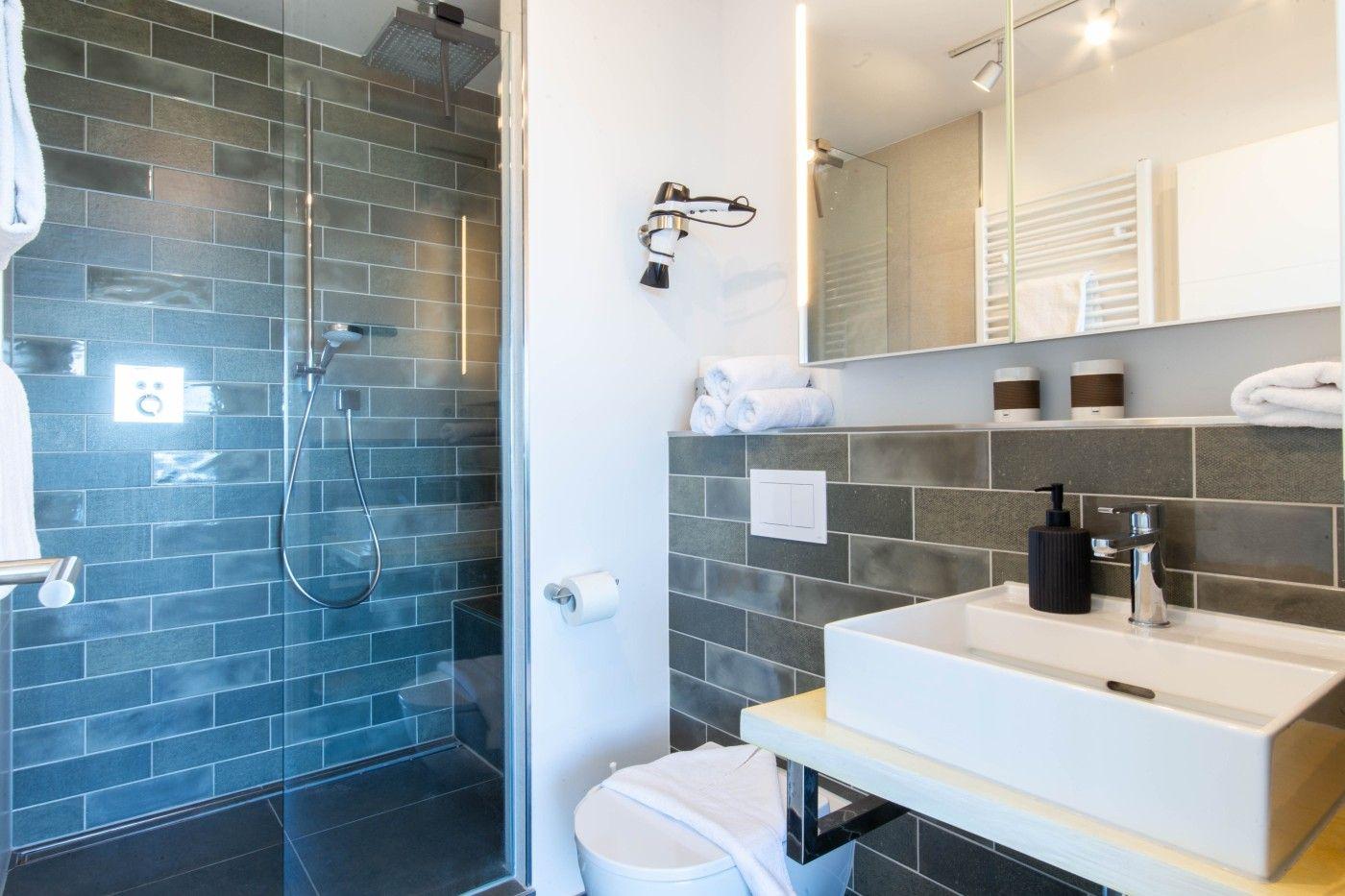 Badezimmer mit Dusche, Waschbecken und Toilette. Fliesenwand und Spiegel sichtbar.