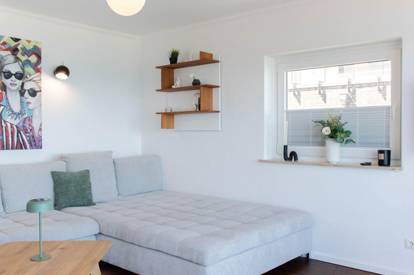 Ein Raum mit Sofa, Wandregal und Fenster mit Blumen.