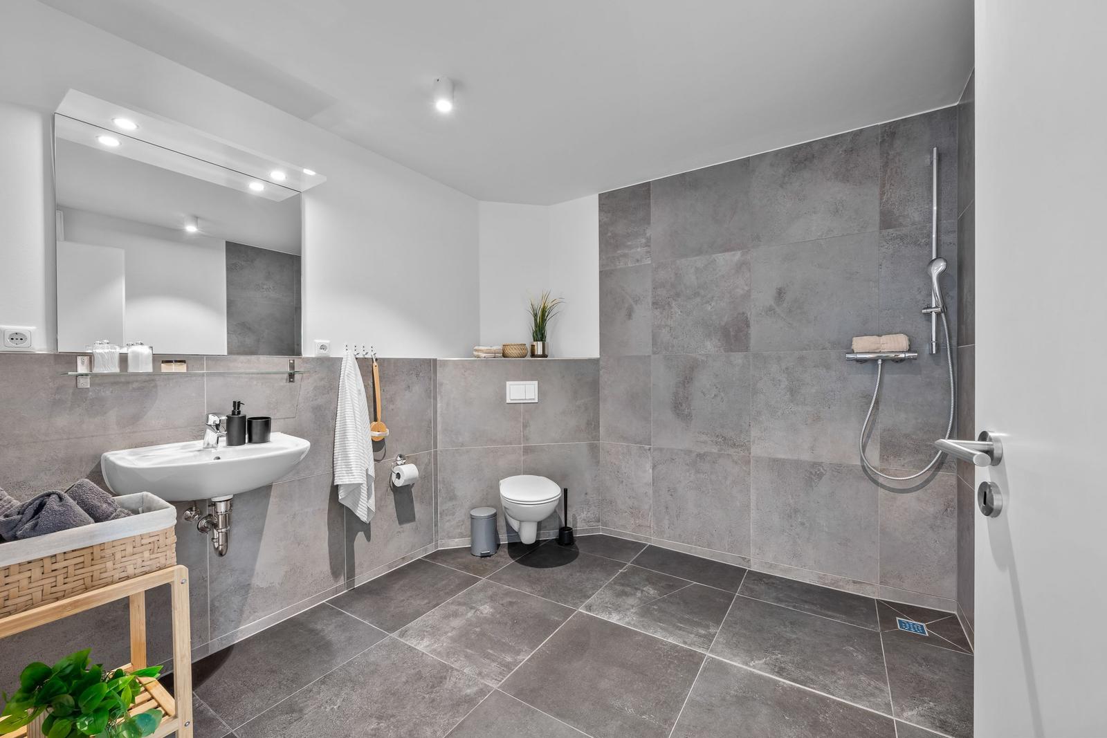 Badezimmer mit Dusche, Waschbecken und Toilette. Graue Fliesen und Spiegelschrank.