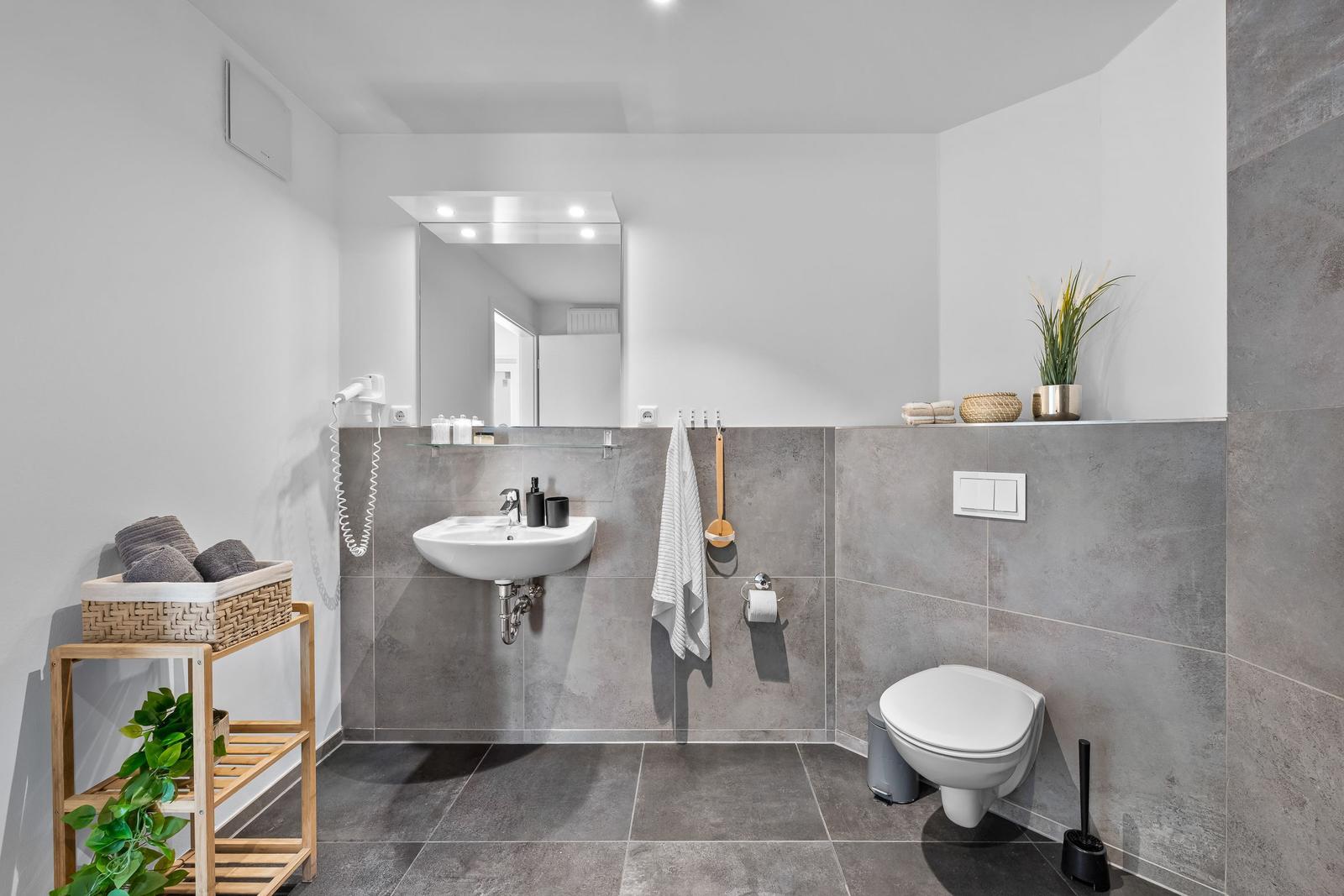 Badezimmer mit Waschbecken, Toilette und Holzregal. Graue Fliesen und Spiegel.