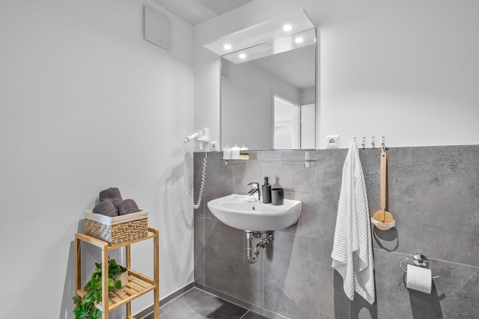 Badezimmer mit Waschbecken, Spiegel, Handtuch und Pflanze auf Holzregal.