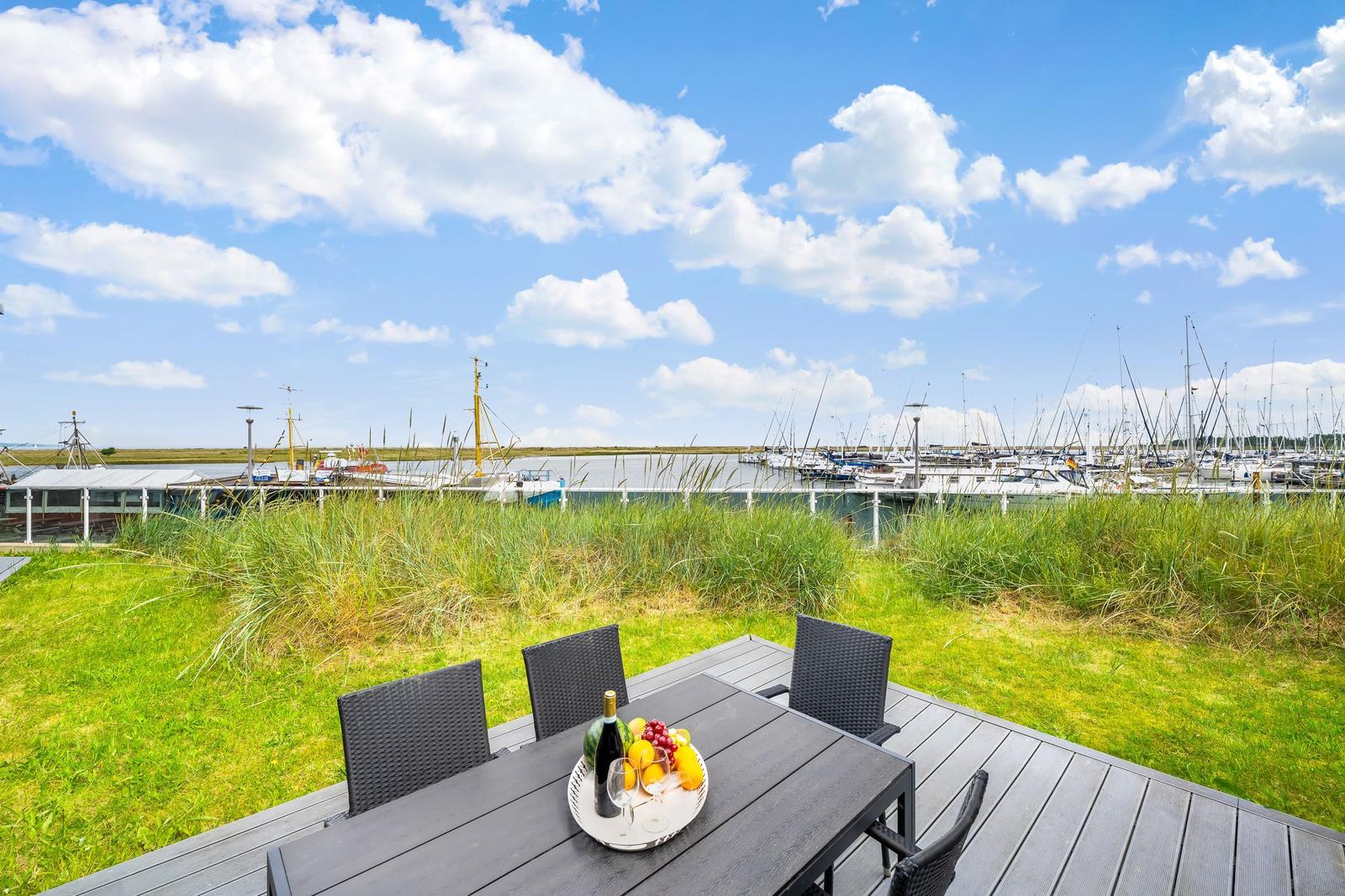 Terrasse mit Tisch und Stühlen, Blick auf Hafen mit Booten und Gras.