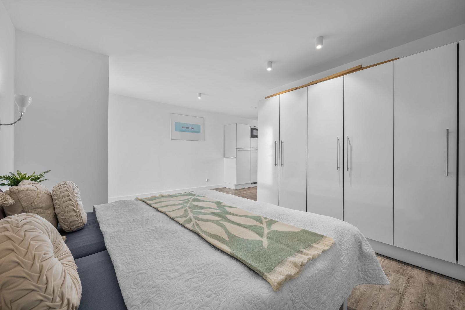 Schlafzimmer mit Bett, Kleiderschrank und Wohnbereich im Hintergrund.