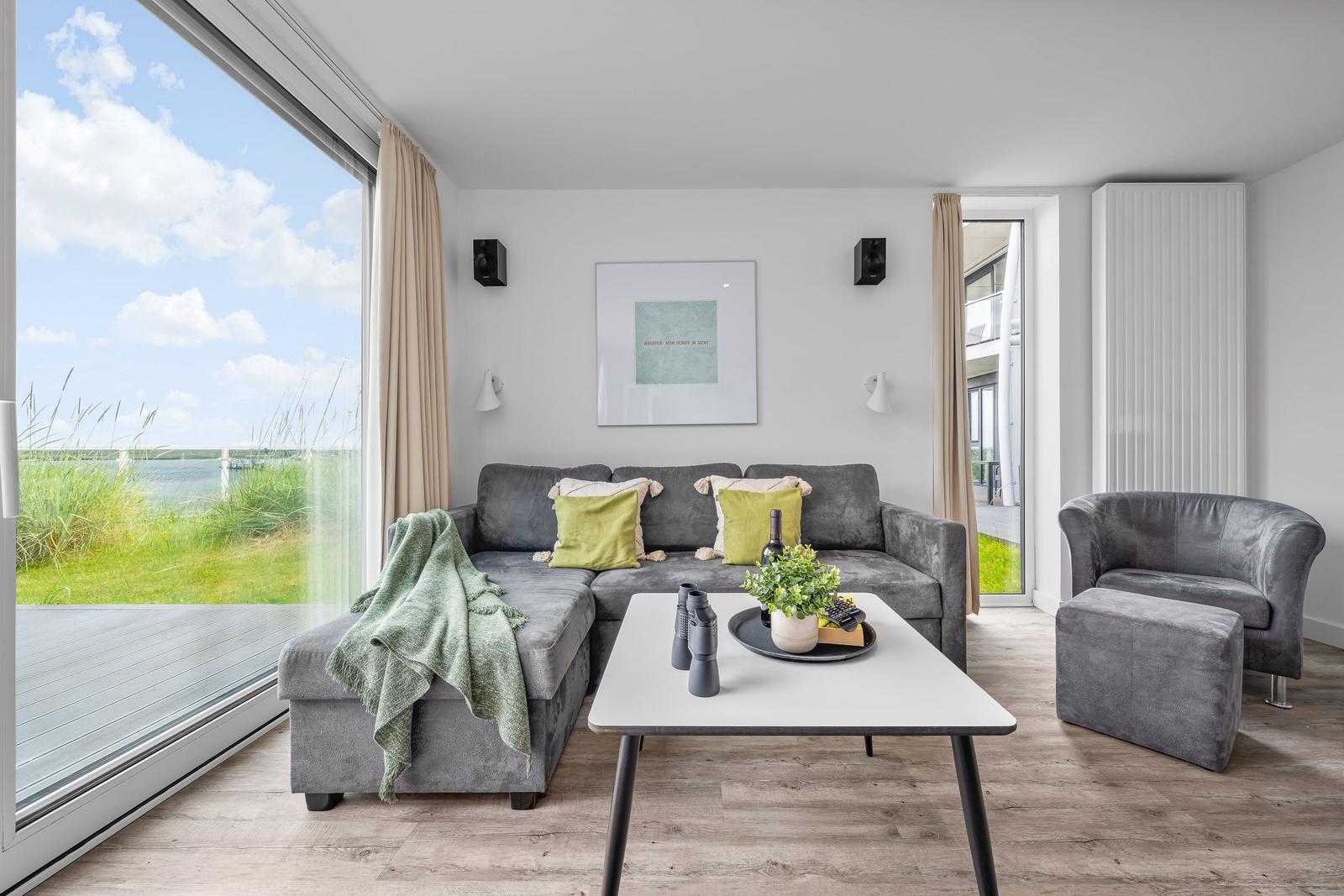Wohnzimmer mit grauem Sofa, Tisch und Blick auf Wasser.