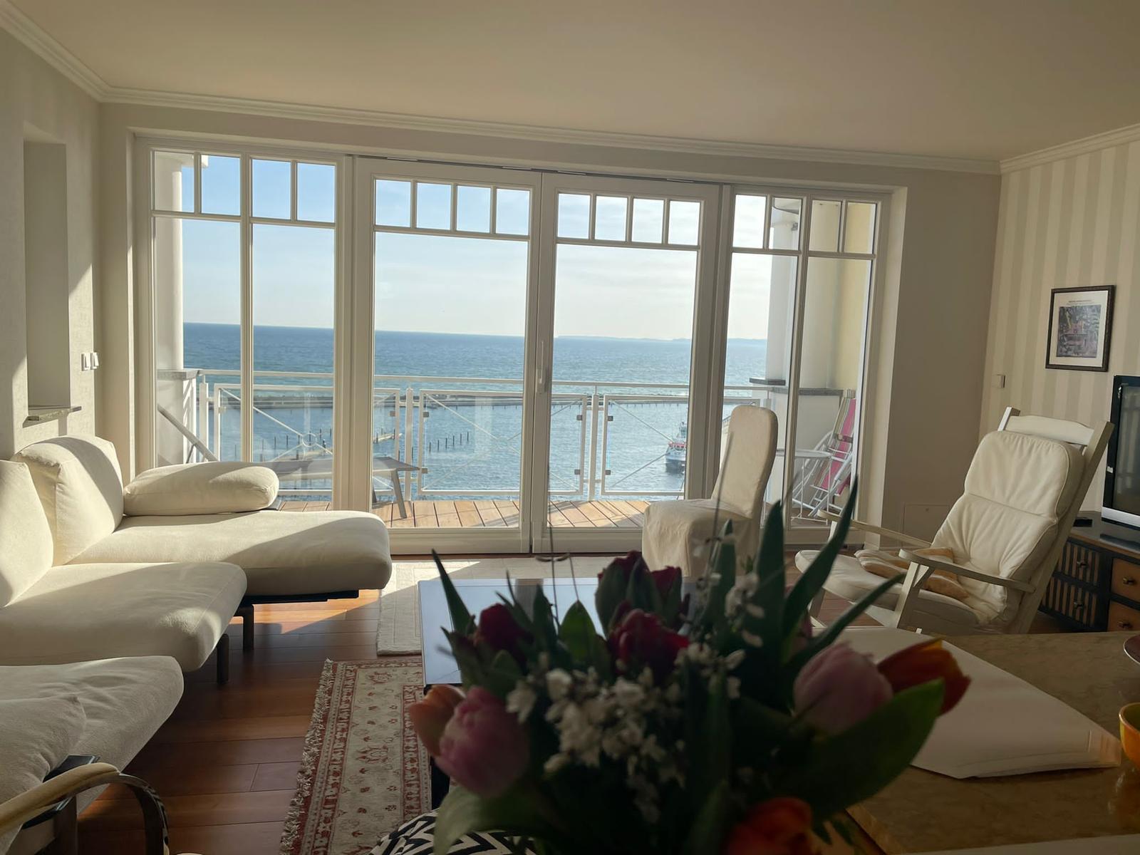 Wohnzimmer mit Meerblick durch große Fenster und Balkon mit Sitzgelegenheiten.