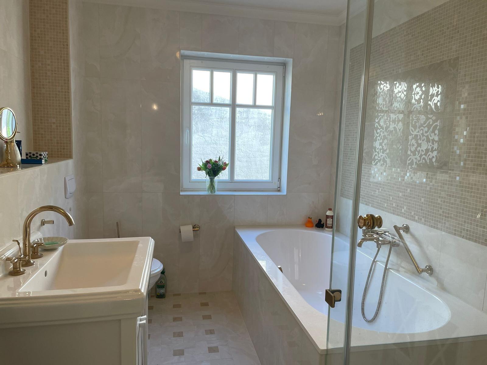 Badezimmer mit Badewanne, Dusche und Waschbecken. Fenster mit Blumen.