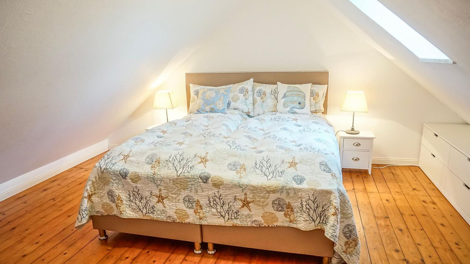 Schlafzimmer mit Bett, Nachttisch und Dachschrägen. Holzboden und Fenster.