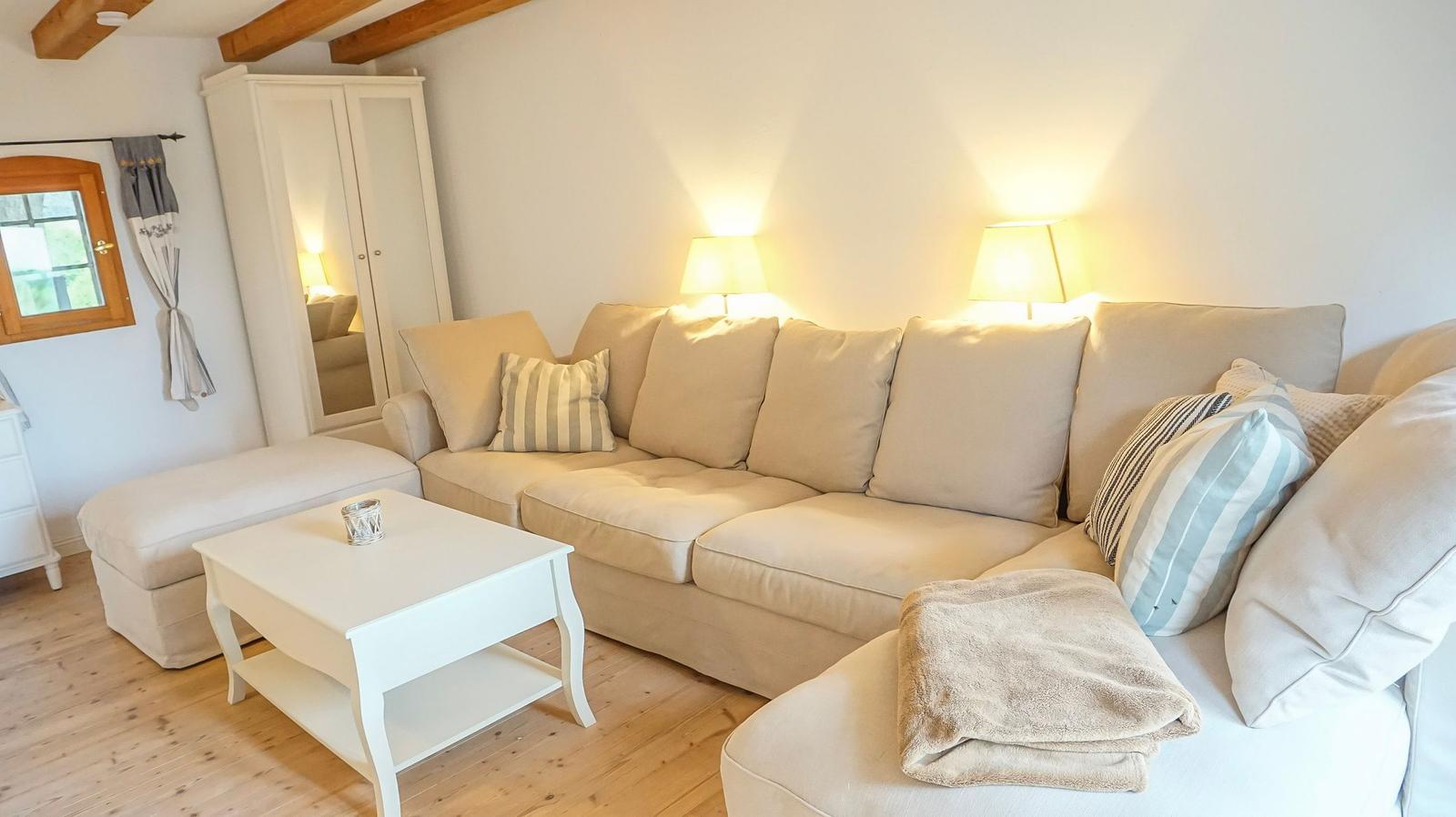 Wohnzimmer mit Ecksofa, Beistelltisch und zwei Stehlampen. Holzboden und weißer Kleiderschrank.