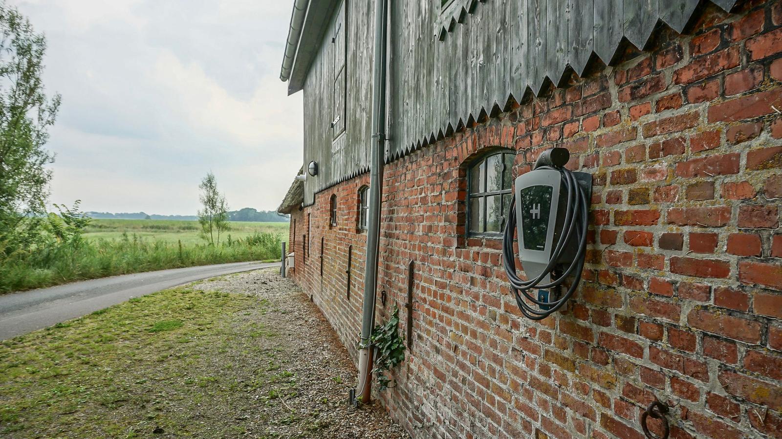 Ladesäule an einer Backsteinmauer neben einem Bauernhof mit Feldweg.
