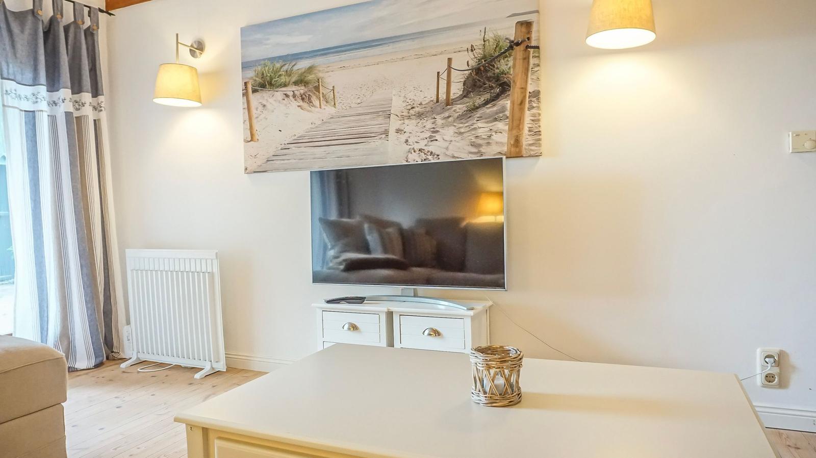 Wohnzimmer mit Fernseher, Wandbild und Tisch. Vorhänge und Heizkörper sind sichtbar.