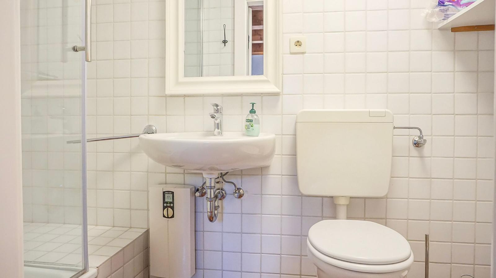 Badezimmer mit Dusche, Waschbecken und Toilette. Weiße Fliesen und Spiegel.