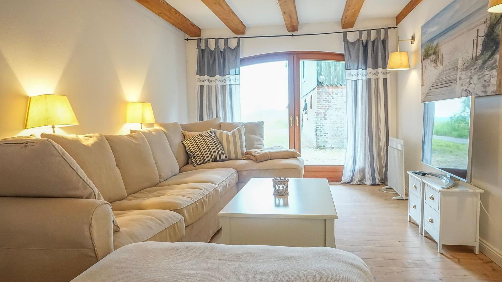 Wohnzimmer mit Sofa, Couchtisch und TV. Holzbalken an der Decke und Fenster mit Vorhängen.