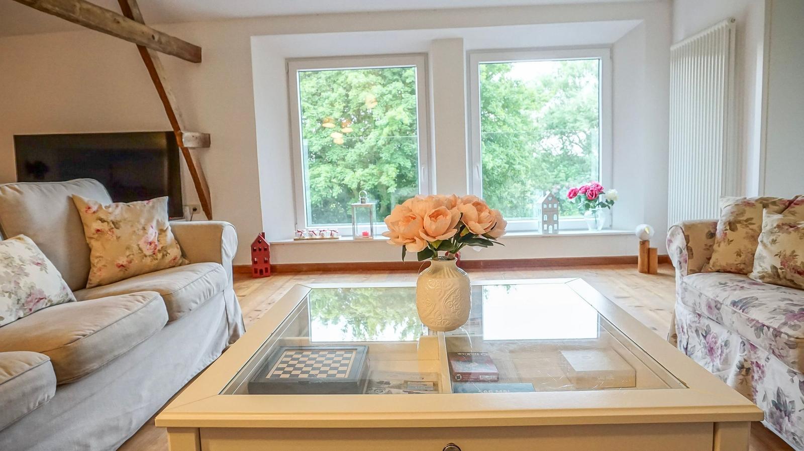 Wohnzimmer mit Sofa, Glas-Tisch und Blick auf grüne Bäume durch Fenster.
