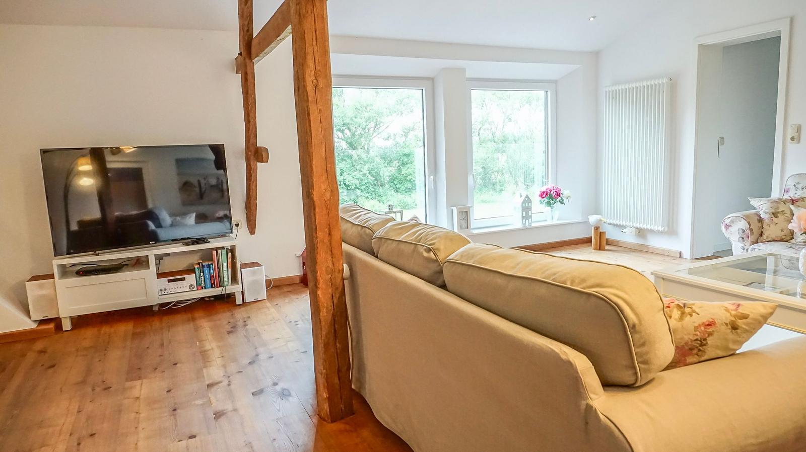 Wohnzimmer mit Sofa, Fernseher und Holzbalken. Große Fenster mit Blick auf Grün.