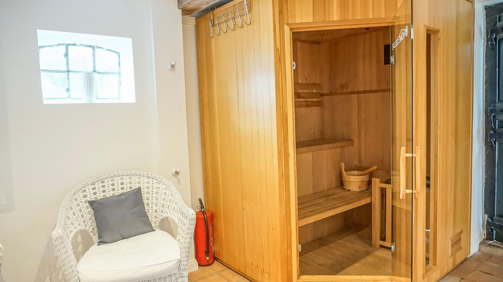 Sauna mit Holzbank und Eimer. Weiße Stuhllehne mit Kissen. Fenster mit Gitter.