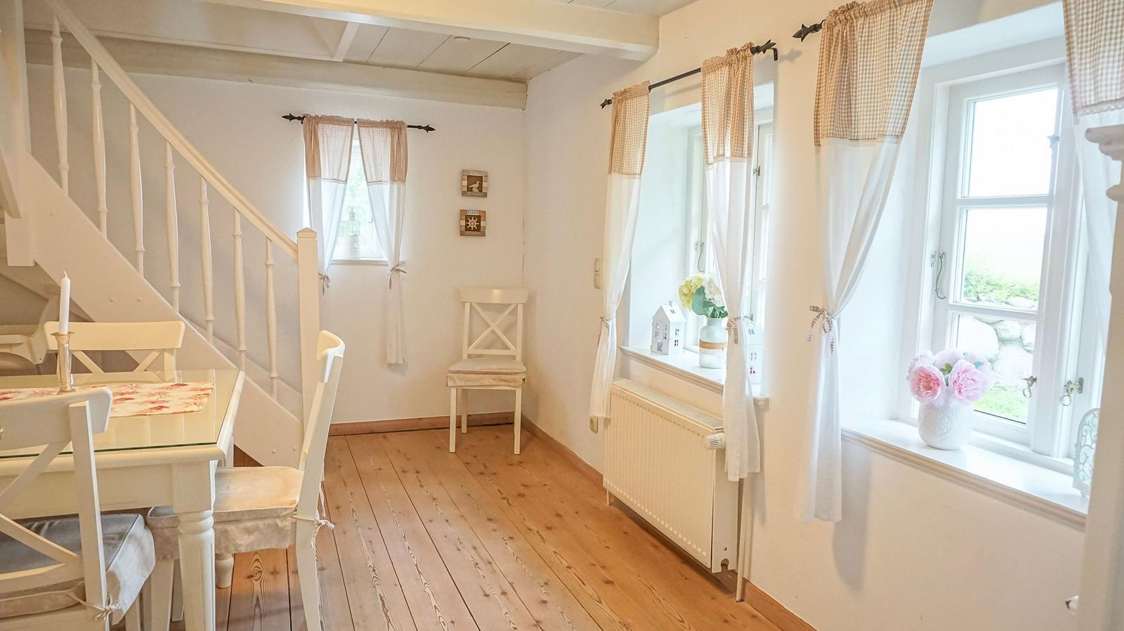 Wohnraum mit Treppe, Tisch, Stühlen und Fenstern mit Vorhängen.