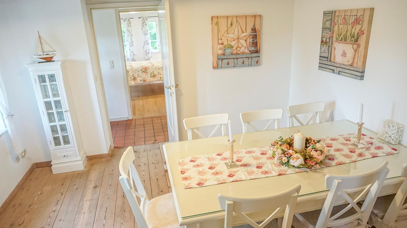 Esszimmer mit Tisch, Stühlen und Wandbildern. Durchgang zu Schlafzimmer mit Bett.