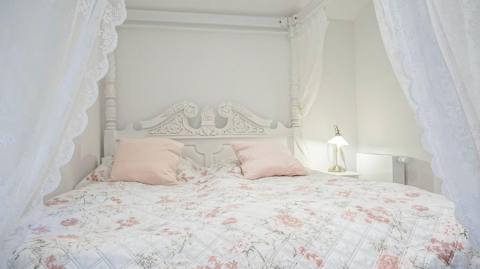 Schlafzimmer mit weißem Bett, floraler Bettwäsche und weißen Vorhängen.