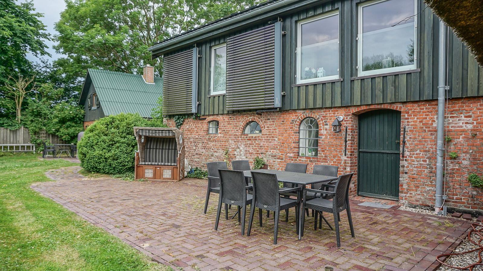 Haus mit Terrasse, Tisch und Stühlen, Beete und Gartenhaus im Hintergrund.