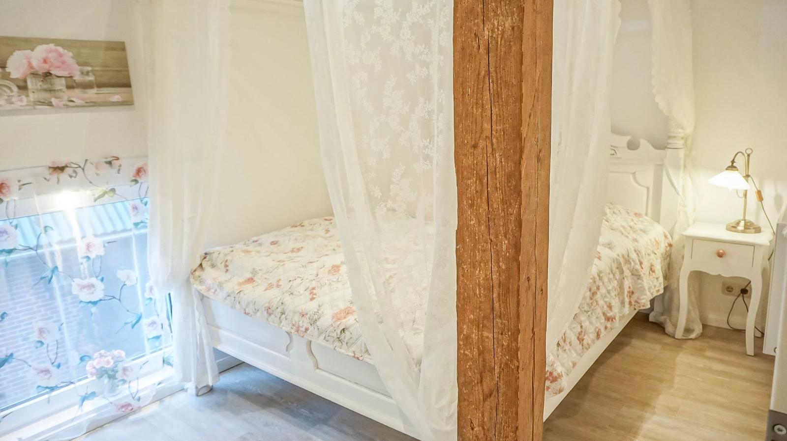 Schlafzimmer mit Bett unter Baldachin, Nachttisch und Fenster mit floraler Gardine.