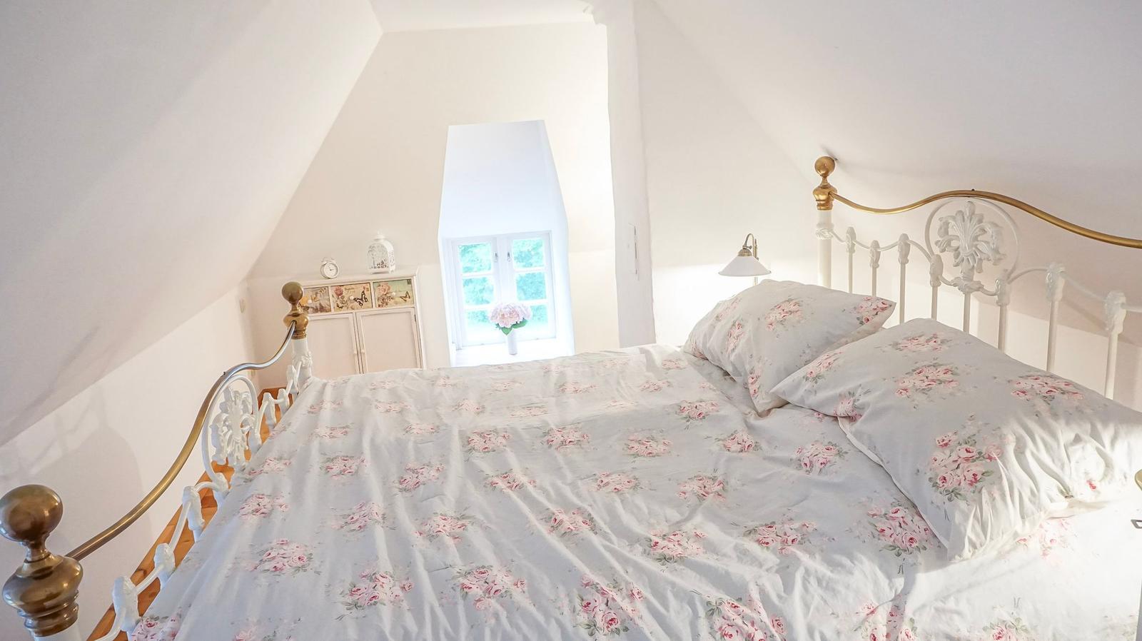 Schlafzimmer mit Dachschrägen, Bett mit Blumenbezug und kleinem Fenster.