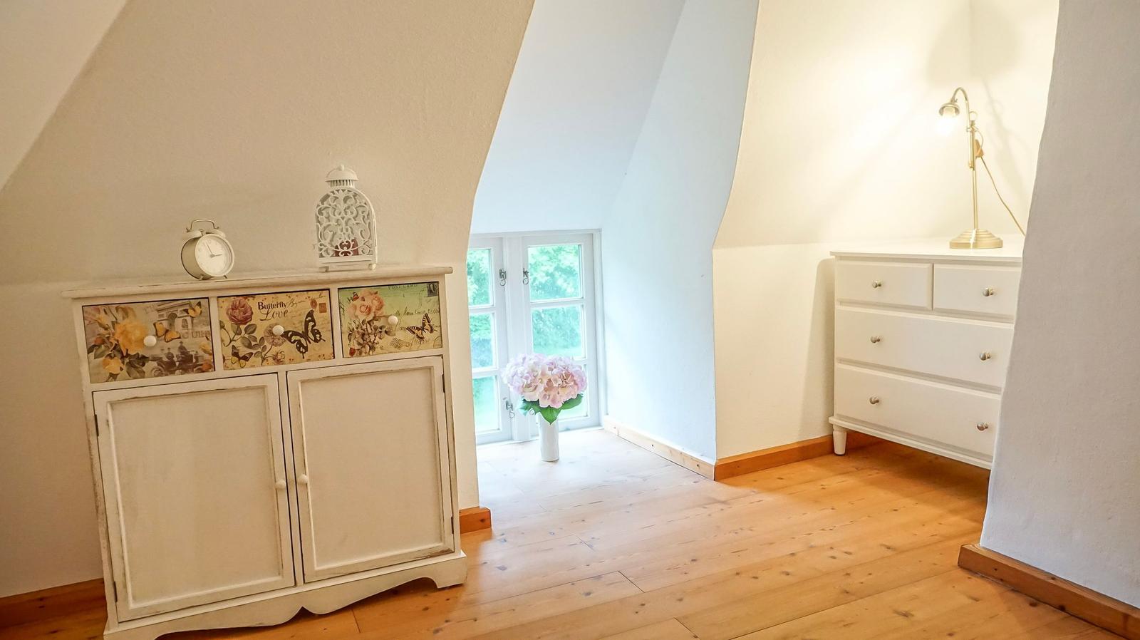 Schlafzimmer mit Holzboden, weißen Schränken und Dachfenster. Auf einem Schrank stehen Wecker und Lampe.