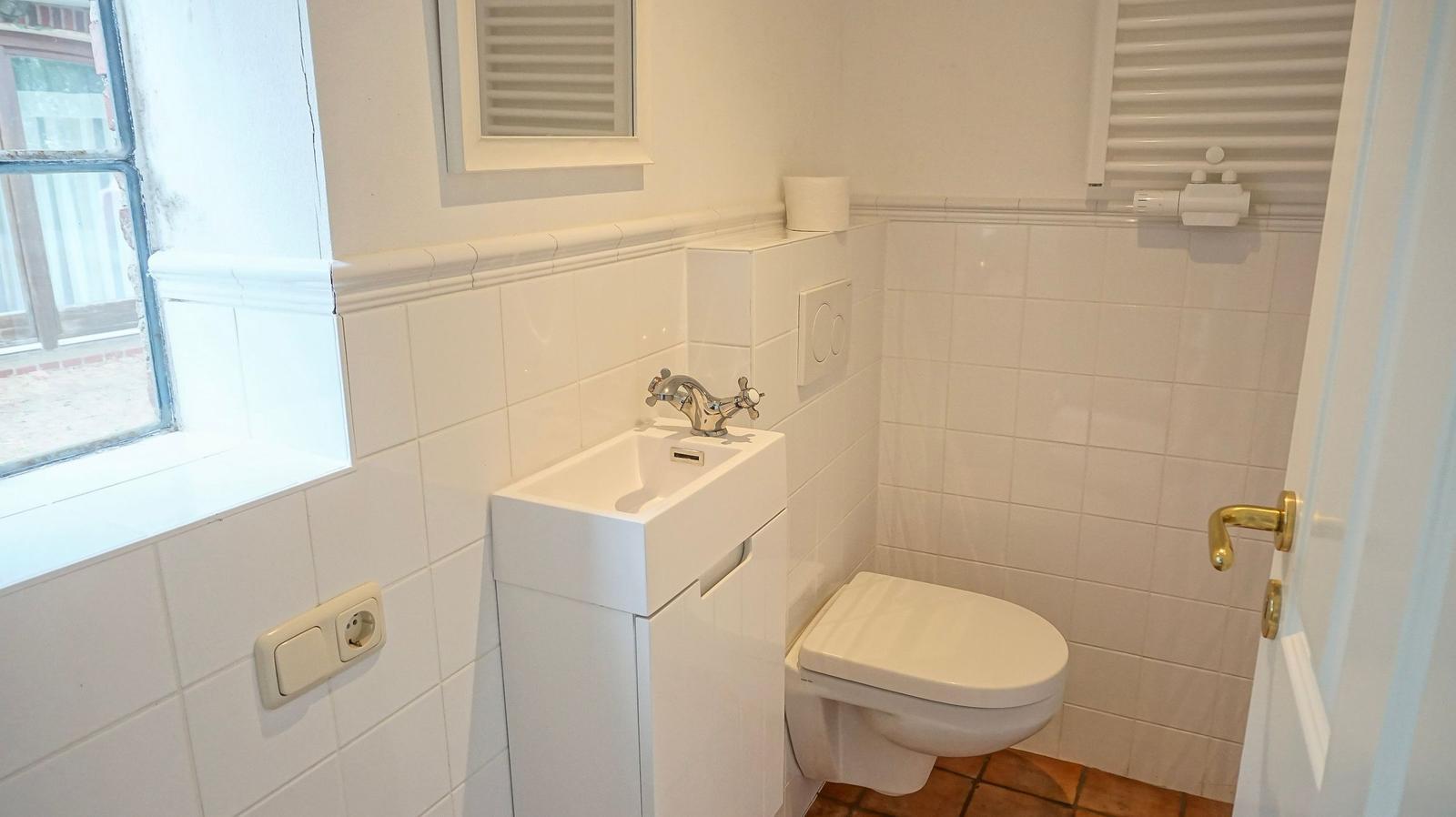 Badezimmer mit Toilette, Waschbecken und Fenster. Wände sind mit Fliesen ausgekleidet.