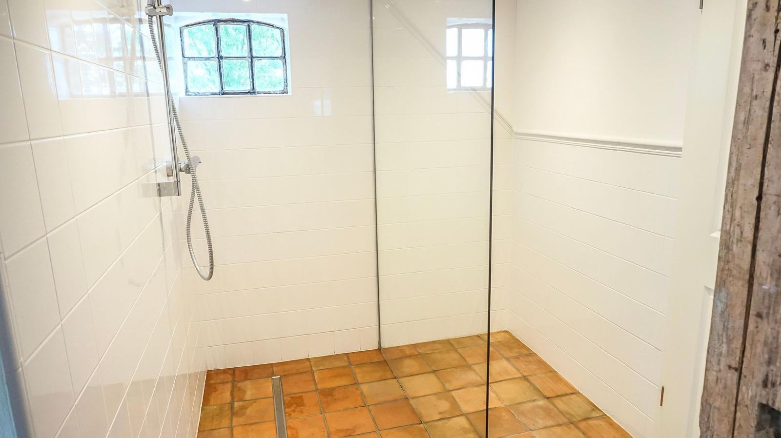 Duschkabine mit Glaswand, weißen Wänden und Terrakottbodenfliesen. Kleines Fenster mit mehreren Glasfeldern.