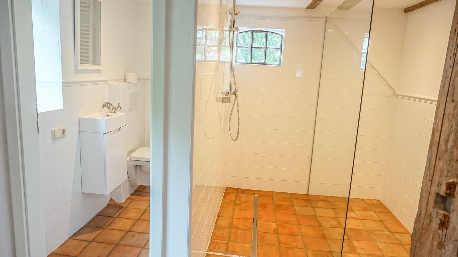 Badezimmer mit Dusche, Toilette und Waschbecken. Terrakottafußboden und Fenster mit Gitter.